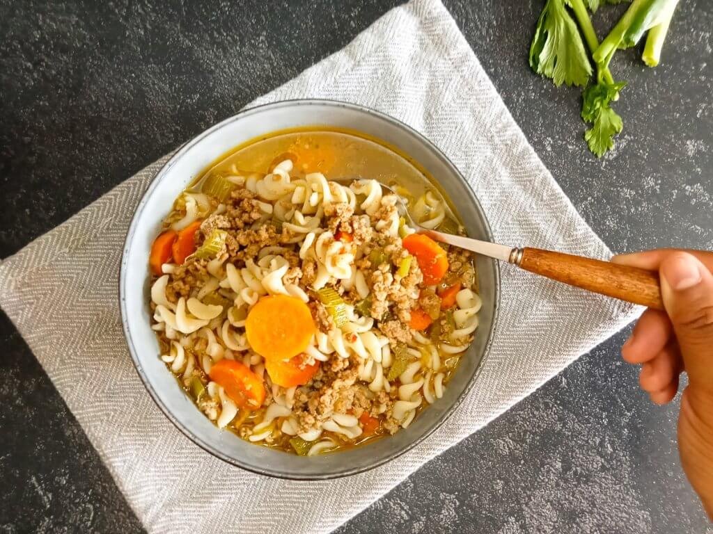 bowl of Vietnamese macaroni soup on a grey table cloth with a human hand scooping a spoonful of the soup with a black mable background.