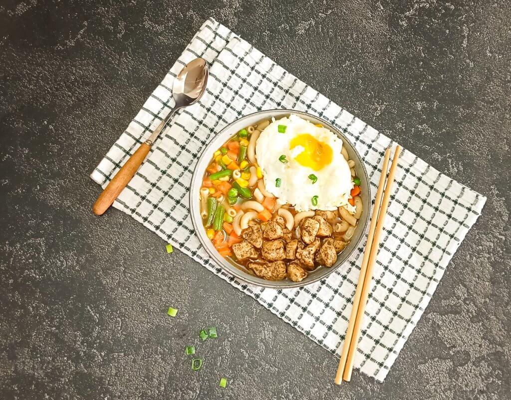 A bowl of Hong Hong 
 style macaroni soup with a spoon beside it as well with chop sticks on a green and white table cloth with a black background.