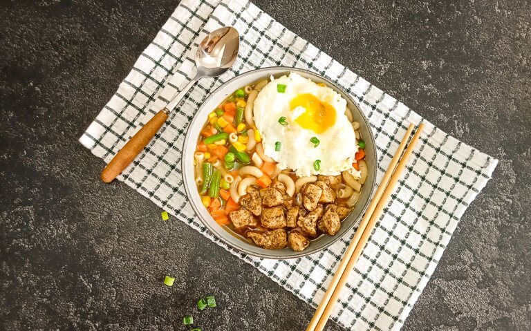 A bowl of Hong Hong macaroni soup with a spoon beside it as well with chop sticks on a green and white table cloth with a black background.