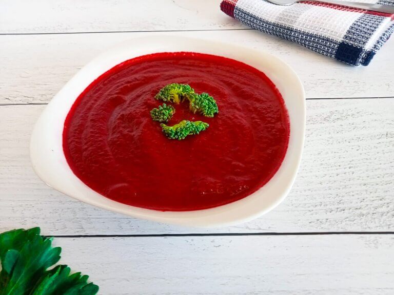 a white bowl containing beetroot and carrot soup on a white pine background with a table cloth and beet greens on the side.