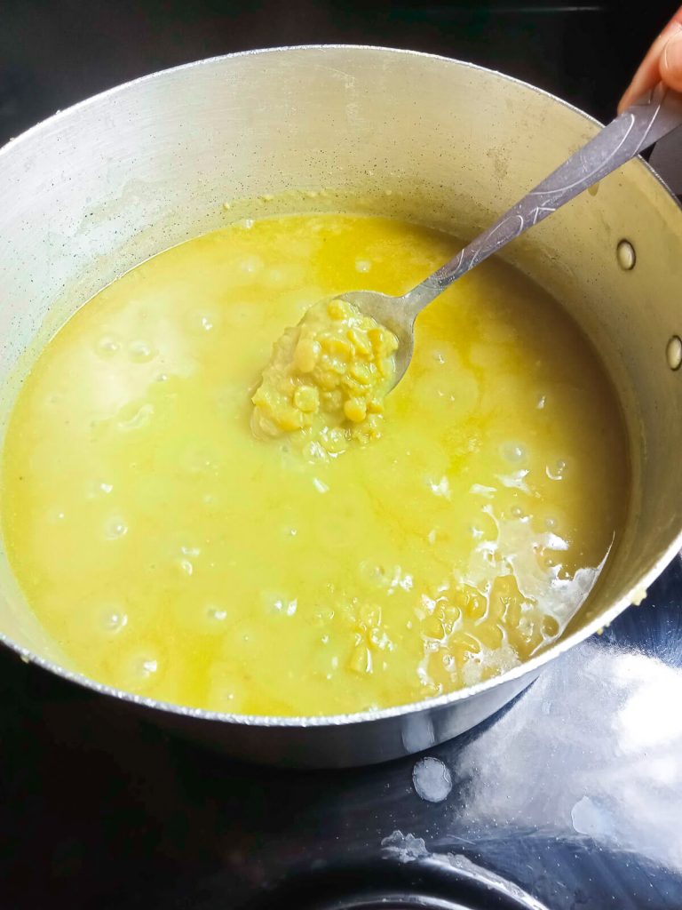 A process photo of a metal pot with green split peas and broth in it and a human hand holding a silver spoon scooping out some split peas.