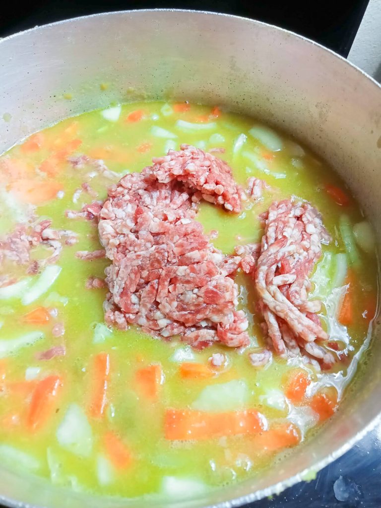 A process photo of a metal pot with onions, carrots, ground beef, and green split peas and broth in it.