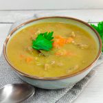 a close up image of a bowl full of green split Pea soup garnished with fresh leaves of herbs on a grey table cloth and withe background.