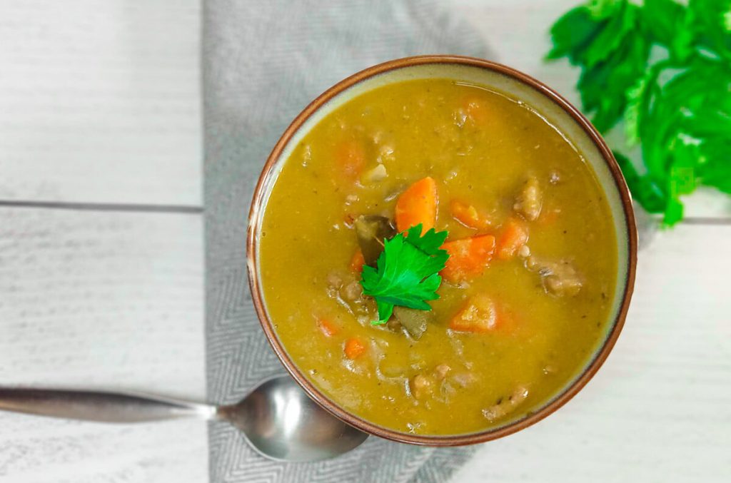 A bowl filled with Goya split Pea soup with a silver spoon and fresh herbs beside it on a grey table cloth and white background.