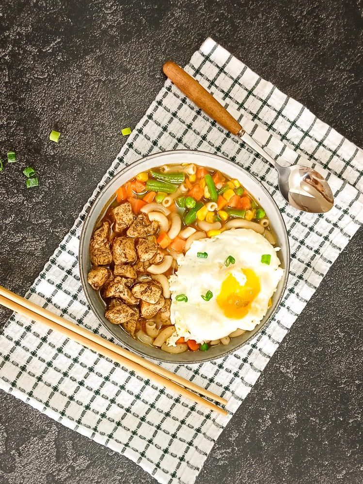 A bowl of Hong Hong macaroni soup with a spoon beside it as well with chop sticks on a green and white table cloth with a black background.