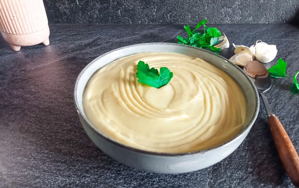 A bowl of mayonnaise soup garnished with fresh leaves of herbs with a spoon beside it and other ingredients on a black background.