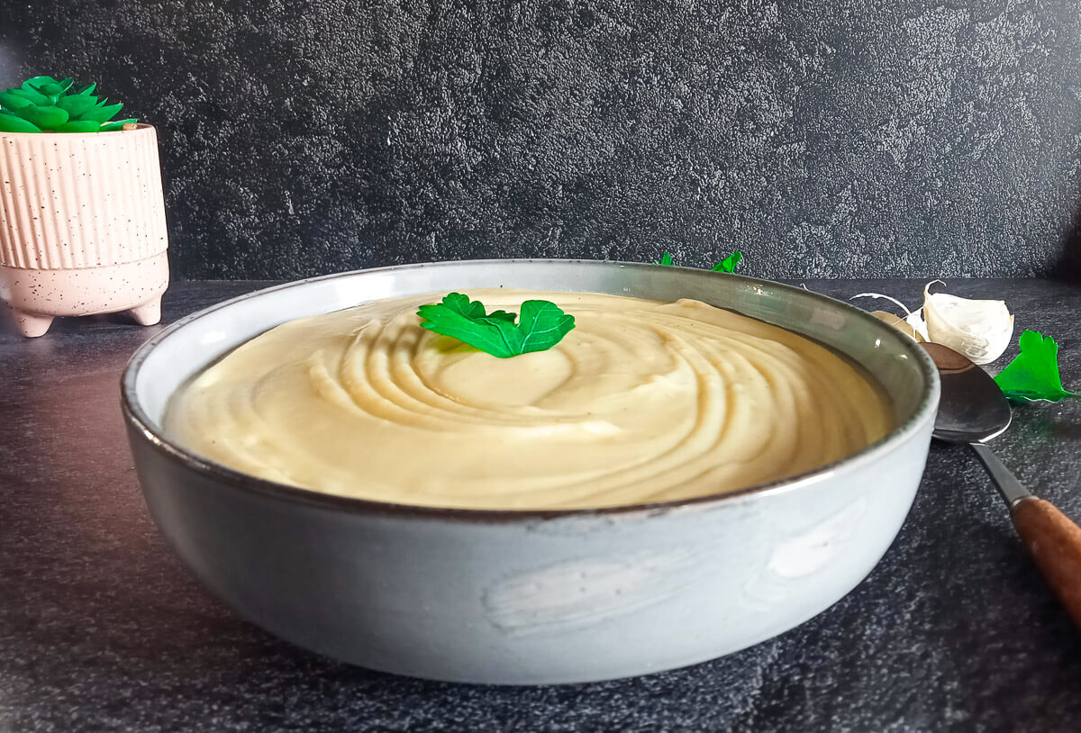 A bowl of mayonnaise soup garnished with fresh leaves of herbs with a spoon beside it and other ingredients on a black background.