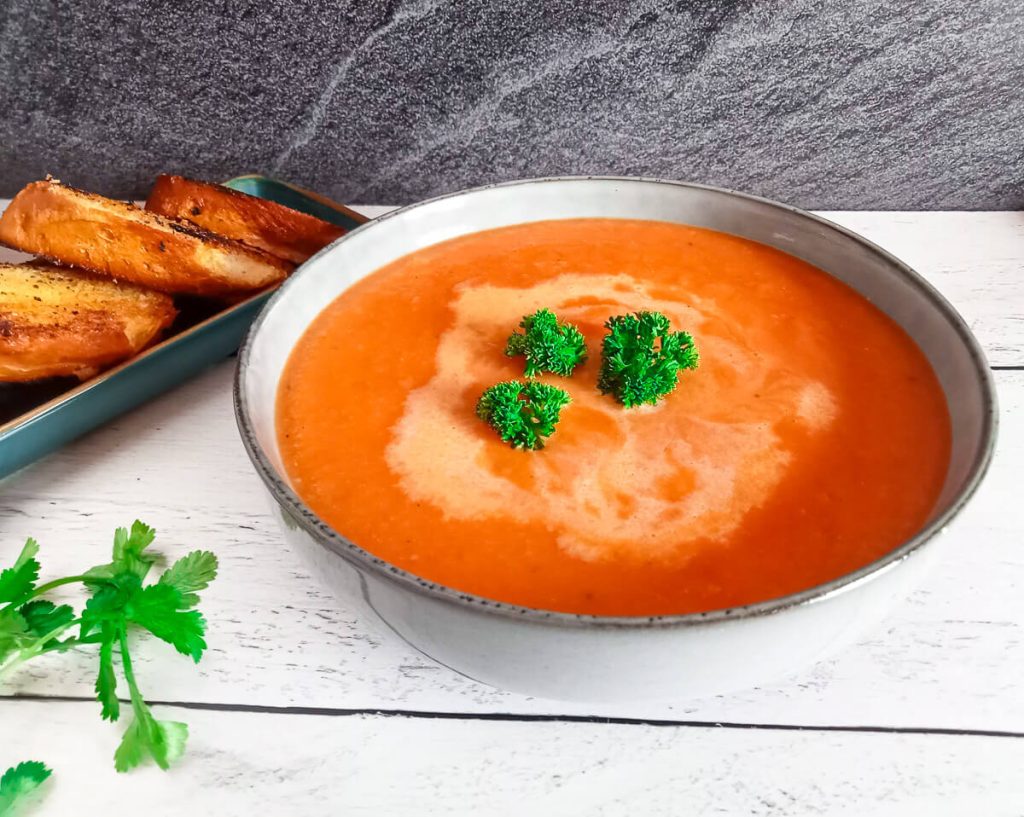 Pepperjack Tomato Soup in a bowl ganished with curly parsley with toasted garlic bread next to the bowl and more fresh herbs scattered on the surface.