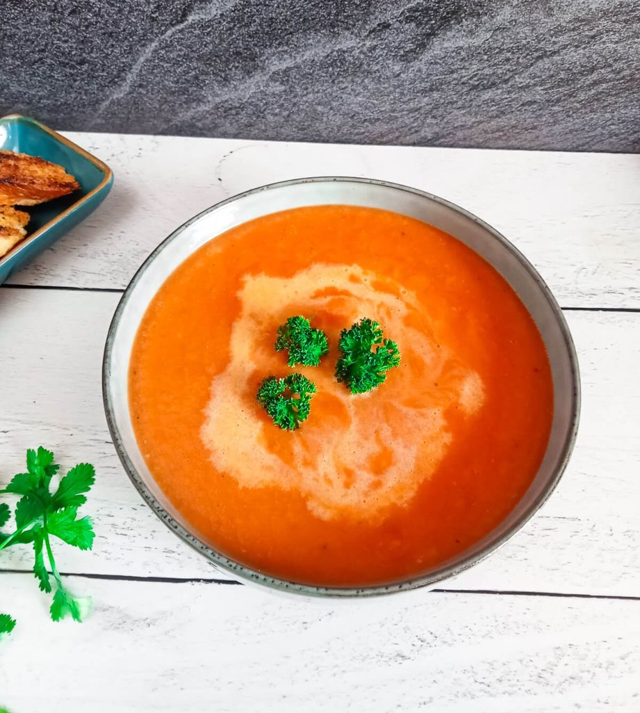 a bowl with Pepper jack Tomato Soup in it with bread and fresh herbs beside it on a white pinewood background.