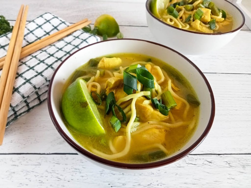 Two bowls of Sukhothai noodle soup with a table cloth and chop sticks on a white background.