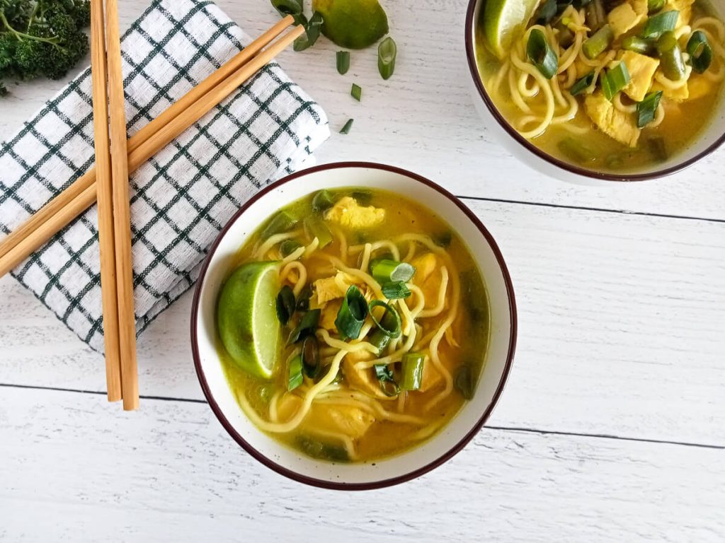 Overhead picture of two bowls of Sukhothai noodle soup with a cut lime and chopped green onion scattered on the surface. there is also a white and green kitchen cloth with chop sticks ontop of it.