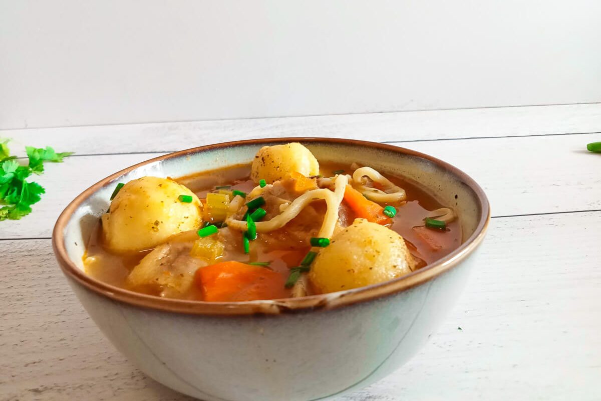 a bowl of mish mash soup which consits of egg noodles, matzo balls , chicken pieces, carrots and broth. This image has a white pinewood background.