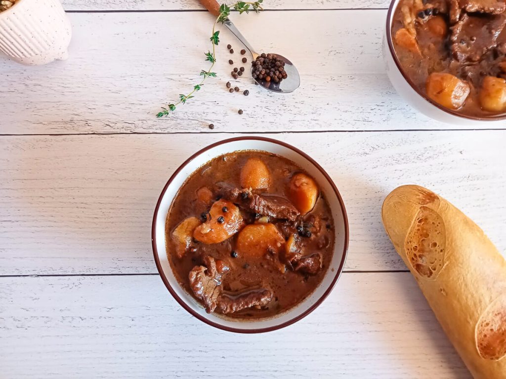 an overhead shot of a bowl filled with steak au poivre soup. Beside it there is black peppercorns in a spoon and dried herbs as well as a loaf of bread on a white pinewood background.