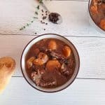 an overhead shot of a bowl filled with steak au poivre soup. Beside it there is black peppercorns in a spoon and dried herbs as well as a loaf of bread on a white pinewood background.