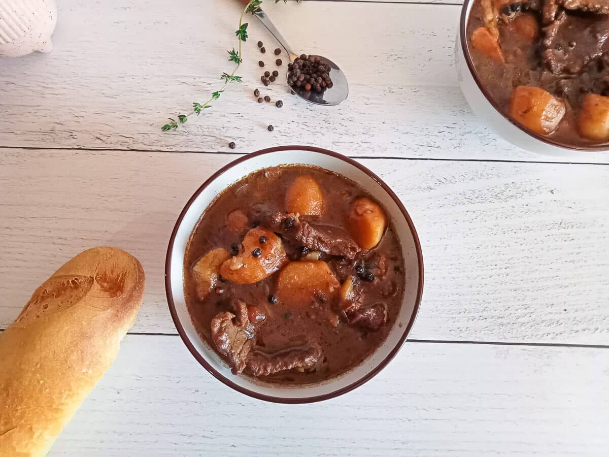 an overhead shot of a bowl filled with steak au poivre soup. Beside it there is black peppercorns in a spoon and dried herbs as well as a loaf of bread on a white pinewood background.