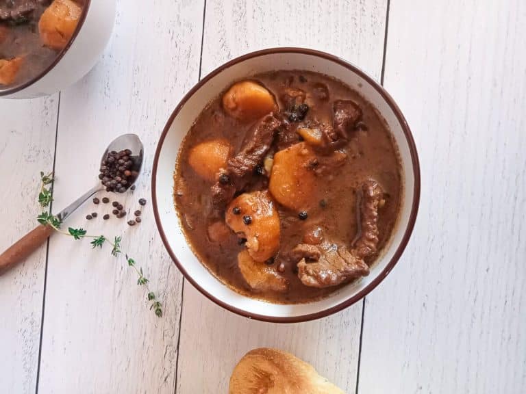 an overhead shot of a bowl filled with steak au poivre soup. Beside it there is black peppercorns in a spoon and dried herbs as well as a loaf of bread on a white pinewood background.