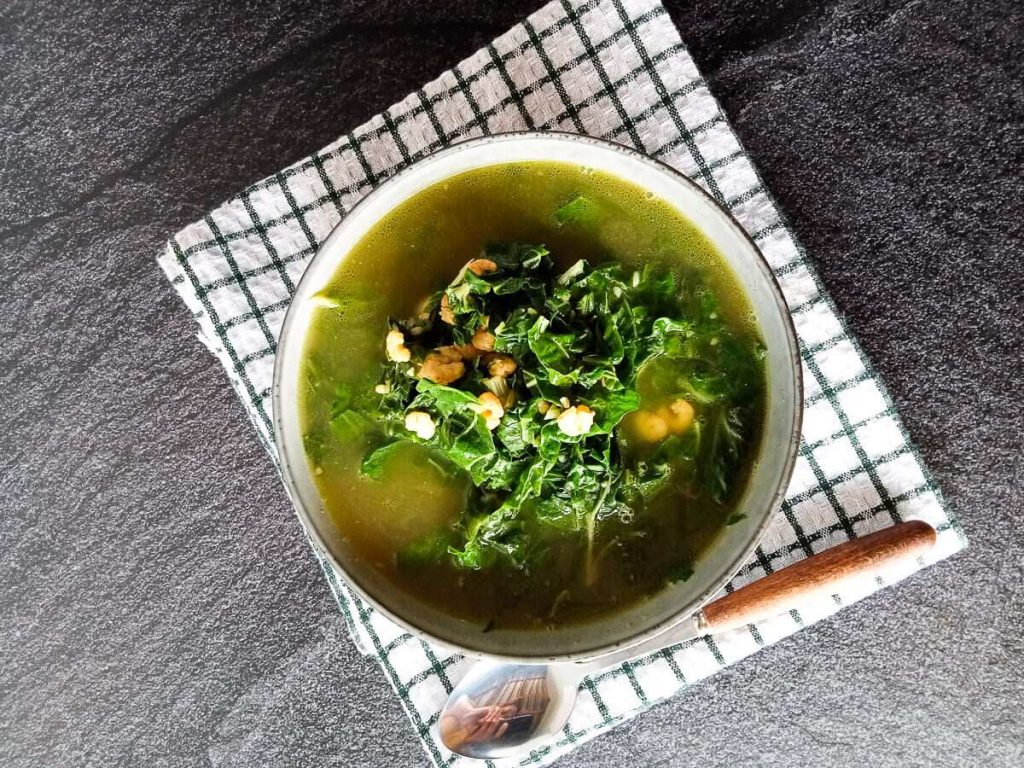 A bowl of Vietnamese spinach soup with shrimp on a folded tablecloth with a silver spoon beside it on a black background.