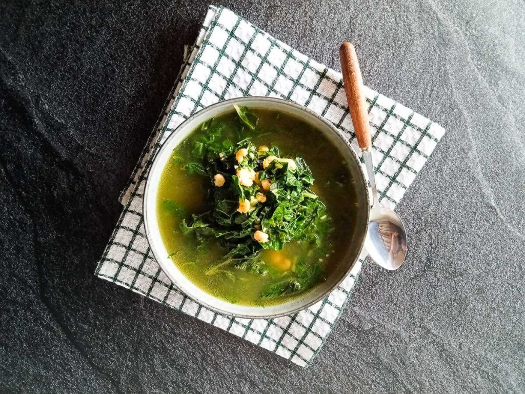 A bowl of Vietnamese spinach soup with shrimp on a folded tablecloth with a silver spoon beside it on a black background.