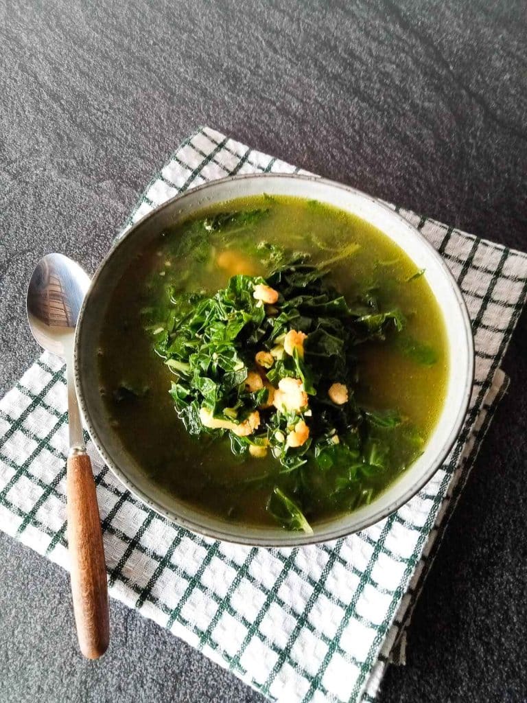 A bowl of Vietnamese spinach soup with shrimp on a folded tablecloth with a silver spoon beside it on a black background.