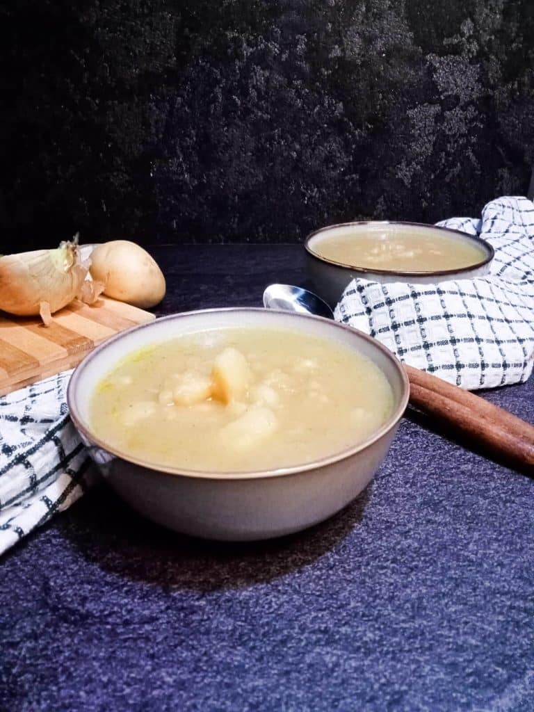 2 bowls of 3 ingredients potato soup with two spoons and a table cloth. There is left over ingredients on a chopping board in the background.