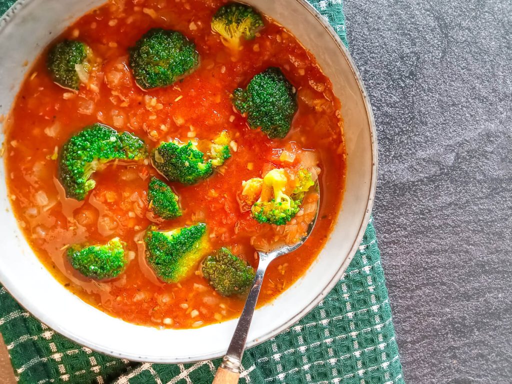 A spoon scooped broccoli tomato soup from a bowl on top of a green and white table cloth with a black marble background.