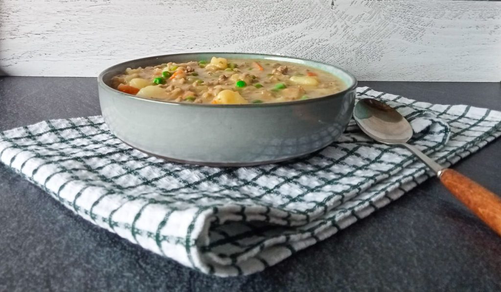 A bowl of potato hamburger soup on a white table cloth with a spoon beside the bowl on a black background.