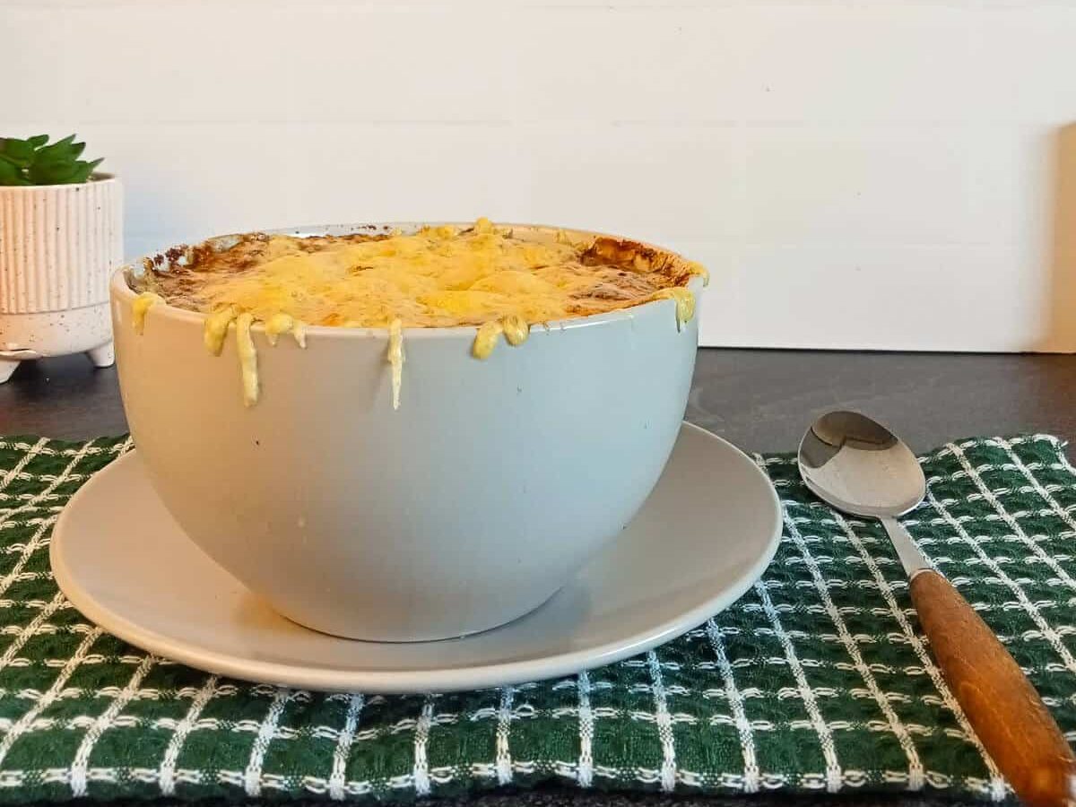 A bowl of Roy Choi french onion soup ontop od a side plate and a green and white table cloth with a silver spoon beside it.