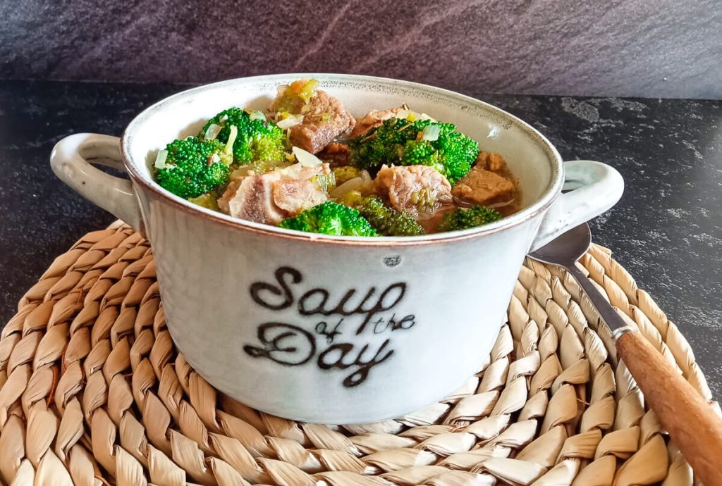 A close-up shot of beef broccoli soup in a bowl on top of a plate mat with a spoon beside it and it is on a black background.