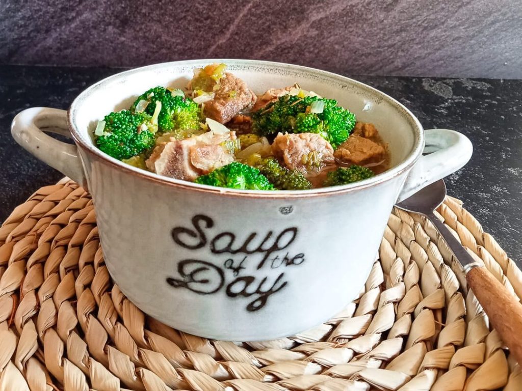 A close-up shot of beef broccoli soup in a bowl on top of a plate mat with a spoon beside it and it is on a black background.