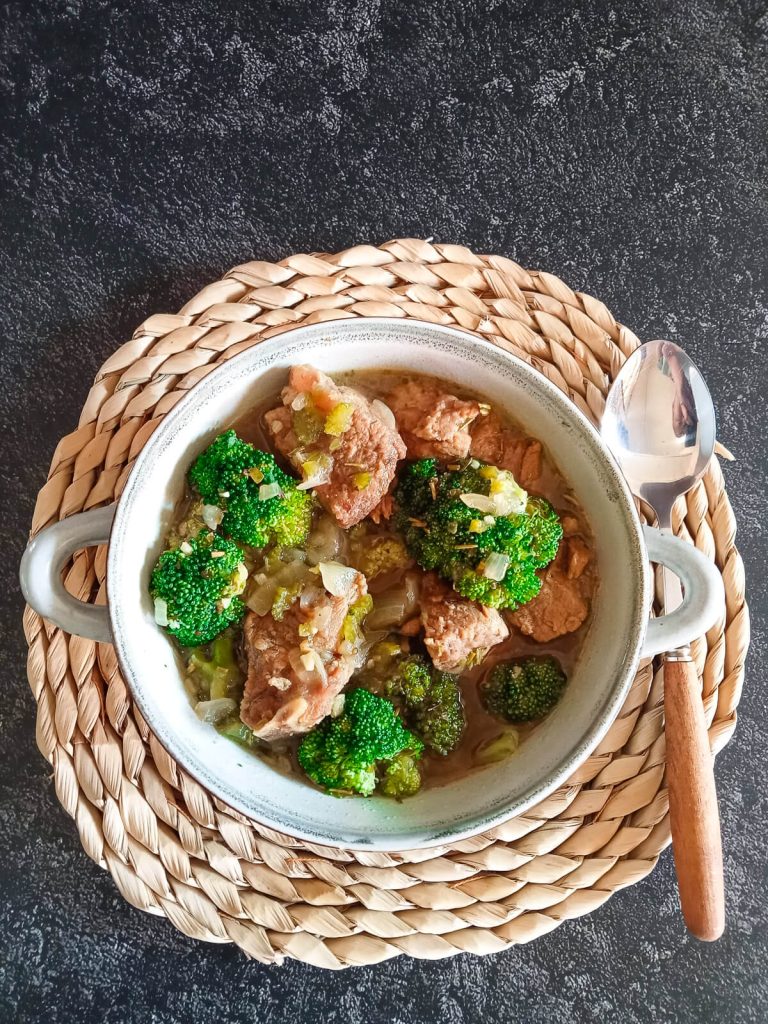 An overhead shot of beef broccoli soup in a bowl on top of a plate mat with a spoon beside it and it is on a black background.