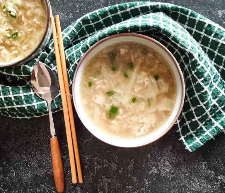 an overhead image of two bowls of egg drop soup with cabbage and 2 chopsticks and a spoon beside the bowl with a green tablecloth on a black background.