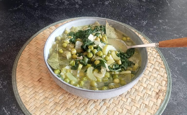 A close-up shot of low-fodmap green minestrone soup in a bowl with a spoon on a plate mat on a black background.