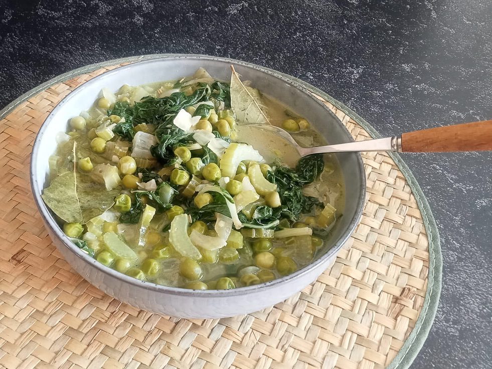 A close-up shot of low-fodmap green minestrone soup in a bowl with a spoon on a plate mat on a black background.