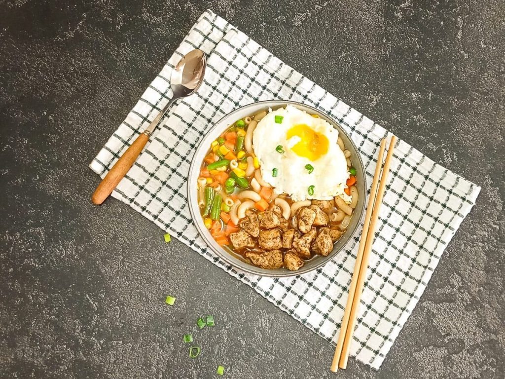 A bowl of Hong Hong style macaroni soup with a spoon beside it as well with chop sticks on a green and white table cloth with a black background.