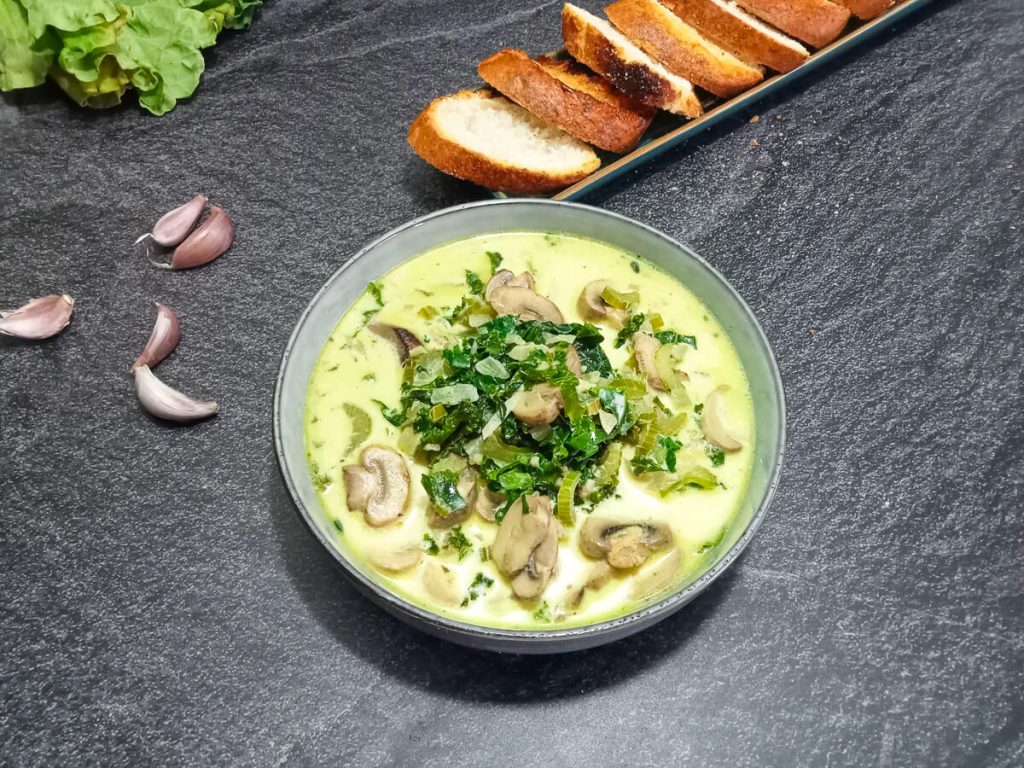 close-up shot of a bowl of kale and mushroom soup with leftover ingredients beside the bowl and sliced garlic bread.