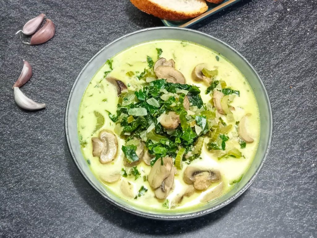 close-up shot of a bowl of kale and mushroom soup with leftover ingredients beside the bowl and sliced garlic bread.