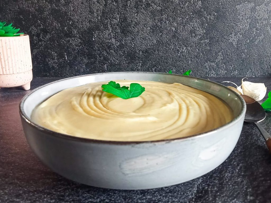 A bowl of mayonnaise soup garnished with fresh leaves of herbs with a spoon beside it and other ingredients on a black background.