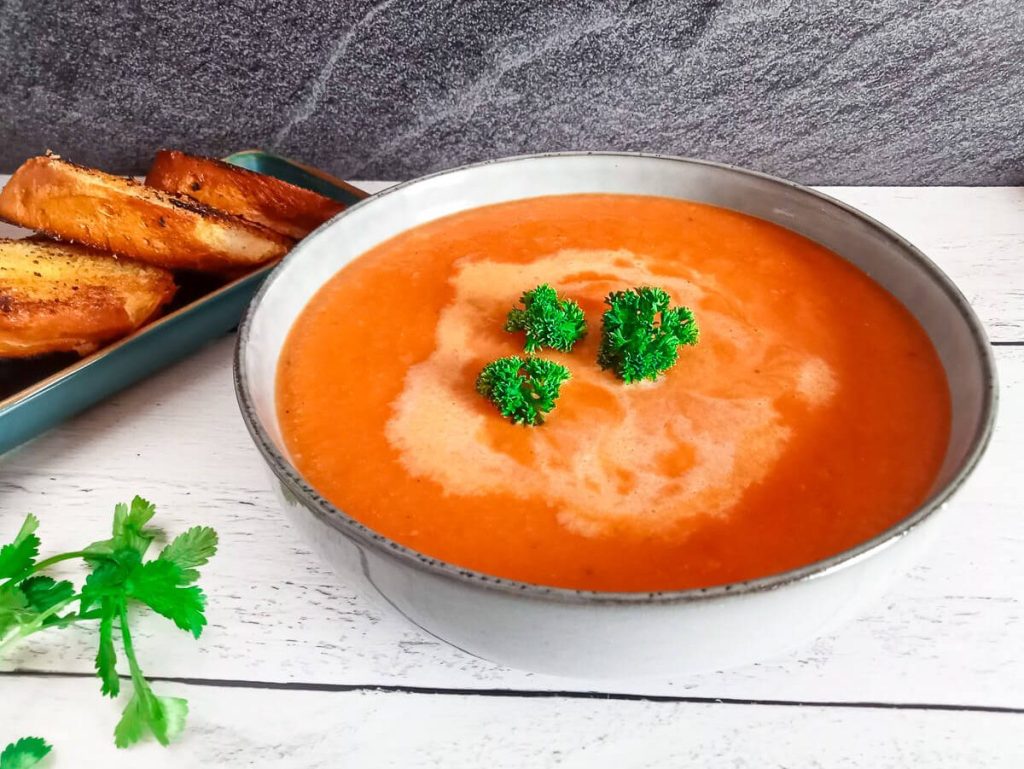 Pepperjack Tomato Soup in a bowl ganished with curly parsley with toasted garlic bread next to the bowl and more fresh herbs scattered on the surface.