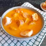 A bowl of Pumpkin chorizo soup with croutons on top and beside the bowl.