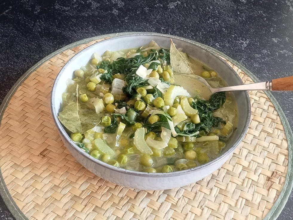 A close-up shot of low-fodmap green minestrone soup in a bowl with a spoon on a plate mat on a black background.