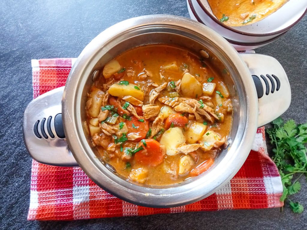 A pot of smoked chicken soup on a tablecloth with serving bowls beside it and fresh herbs