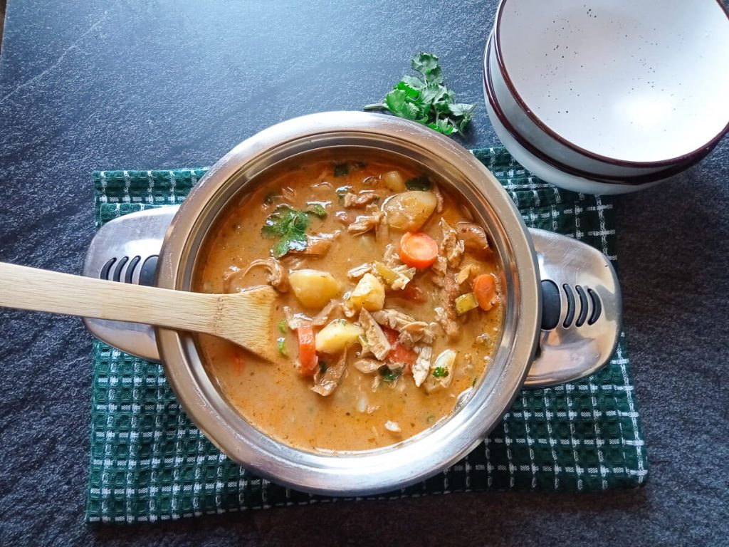 An overhead shot of a pot with marry me chicken soup in it with a wooden spoon. There are empty serving bowls beside the pot which is on top of a green table cloth with fresh herbs beside it all this on a black background.