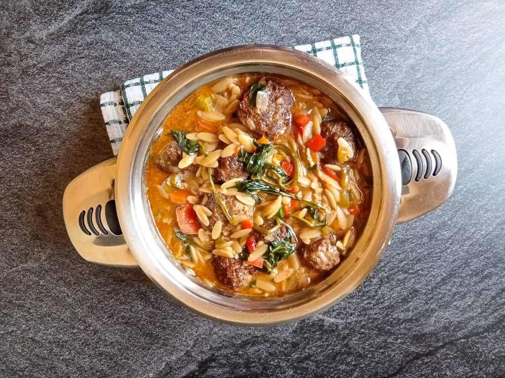 Overhead shot of a silver pot with orzo meatball soup in it on a tablecloth on a black background.