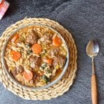 Overhead shot of a bowl of orzo meatball soup on a plate mat with a spoon beside it and a tablecloth in the corner on a black background.