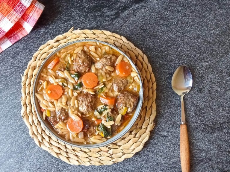 Overhead shot of a bowl of orzo meatball soup on a plate mat with a spoon beside it and a tablecloth in the corner on a black background.