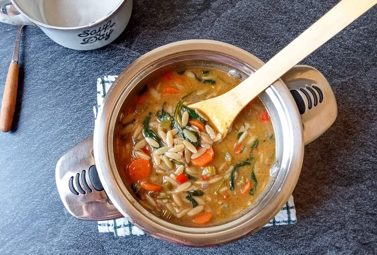 An overhead shot of a pot full of vegetable orzo soup with a wooden spoon on a tablecloth. Beside it there is an empty bowl and a silver spoon