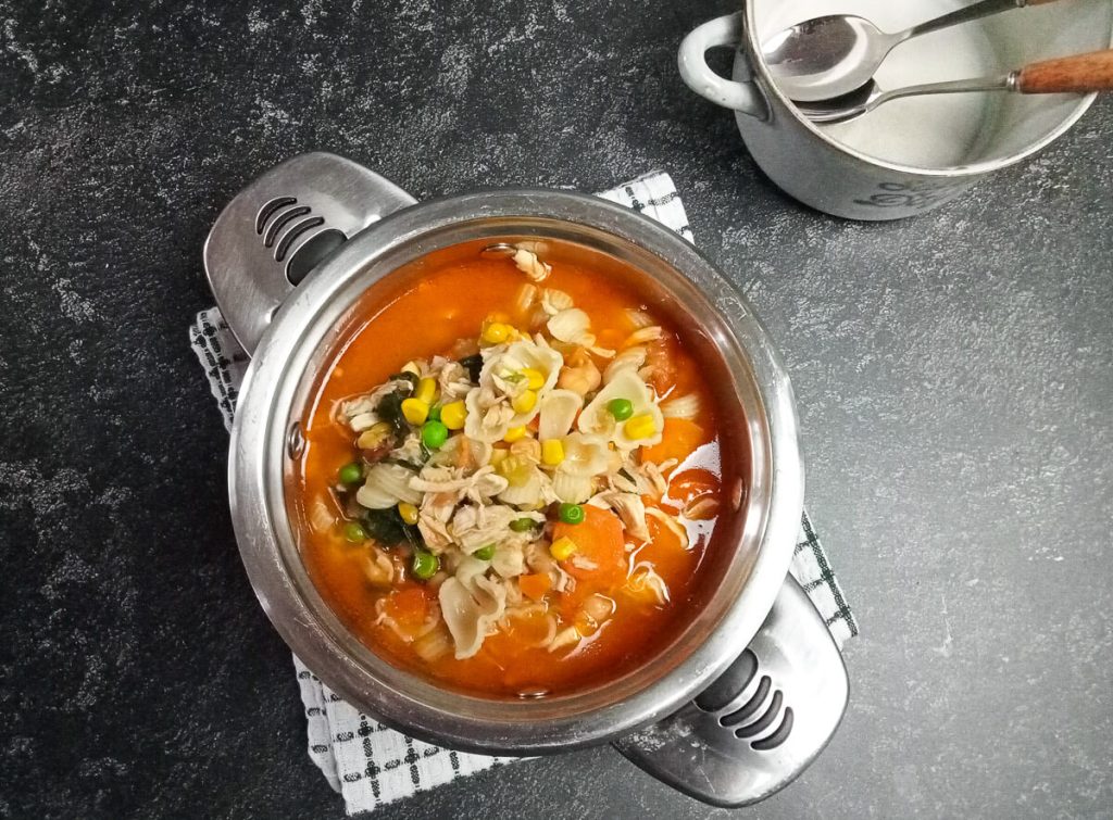 A shot of high-protein chicken noodles soup in a pot on top of a white and green tablecloth with an empty serving bowl beside it