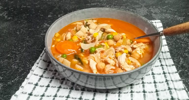 A shot of high-protein chicken noodles soup in a bowl on top of a white and green tablecloth with a black background