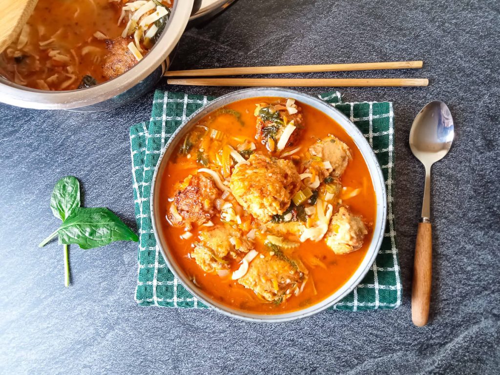 A bowl of Thai chicken meatball soup on a green tablecloth with spoon and chopstick beside it.