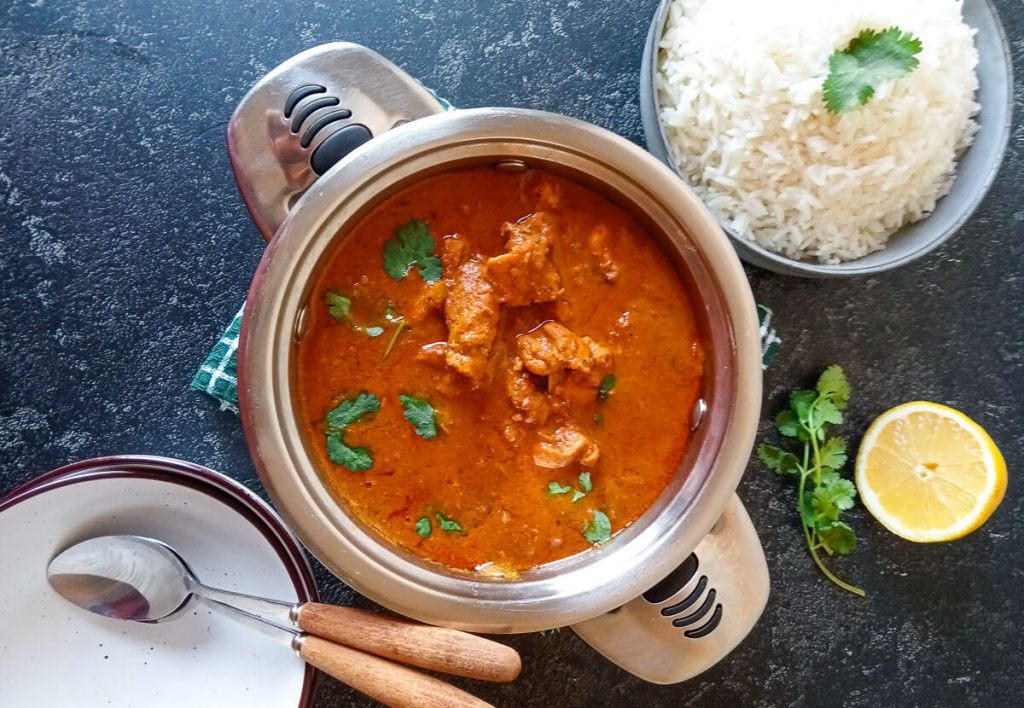 An overhead shot of tikka chicken masala soup in a pot with empty bowls beside it with another bowl of rice.